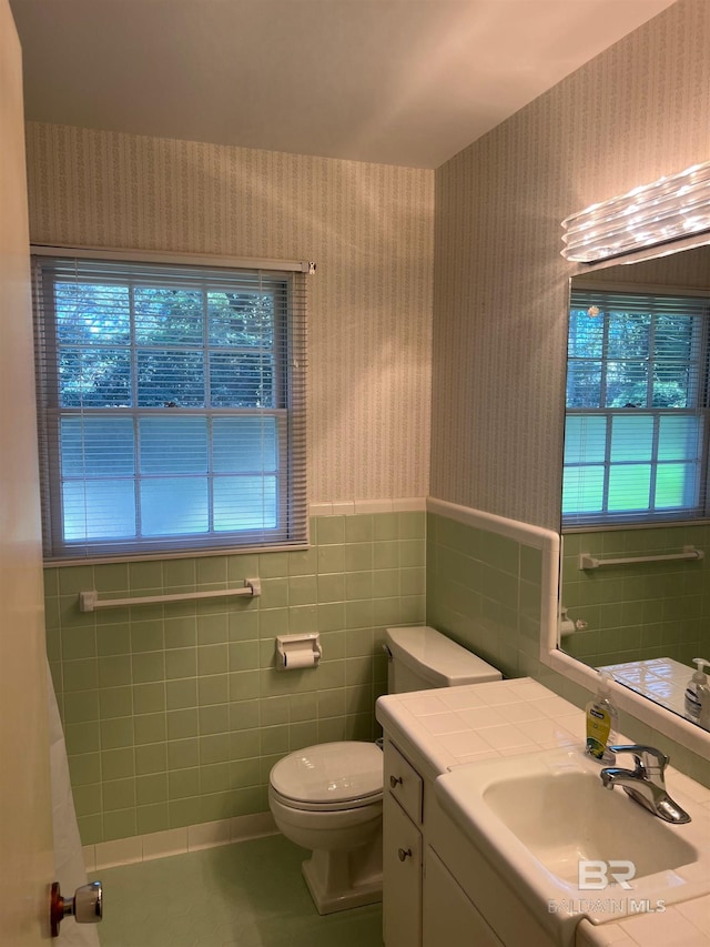 bathroom featuring tile walls, toilet, wainscoting, vanity, and wallpapered walls