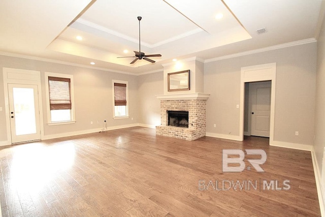 unfurnished living room featuring hardwood / wood-style floors, a brick fireplace, ceiling fan, ornamental molding, and a tray ceiling