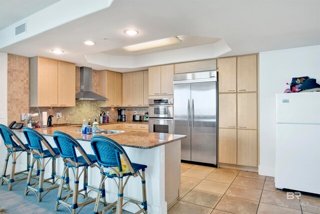 kitchen with kitchen peninsula, light brown cabinetry, stainless steel appliances, and wall chimney range hood