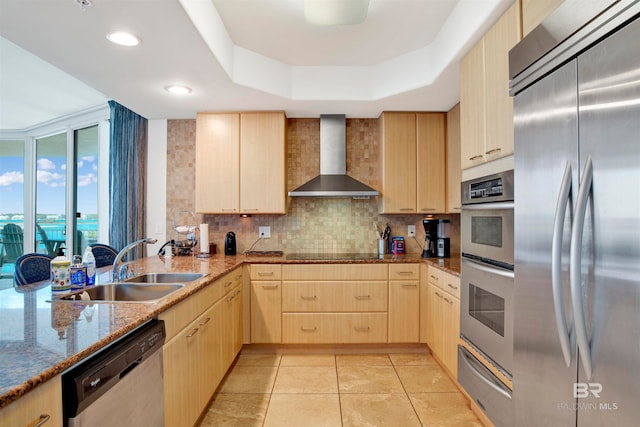 kitchen with light brown cabinets, sink, stainless steel appliances, wall chimney range hood, and a raised ceiling