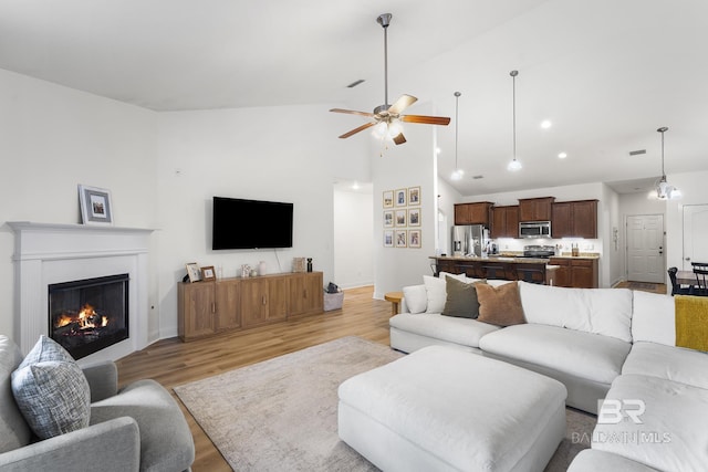 living room with baseboards, light wood-style flooring, a glass covered fireplace, high vaulted ceiling, and a ceiling fan