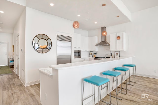 kitchen with white cabinetry, wall chimney exhaust hood, kitchen peninsula, built in appliances, and decorative light fixtures