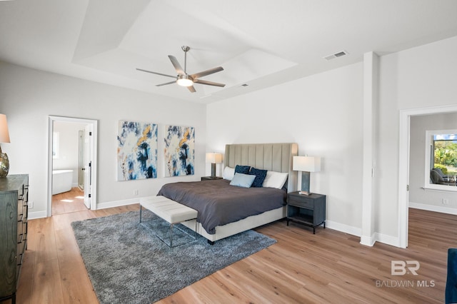 bedroom with ceiling fan, wood-type flooring, a tray ceiling, and ensuite bath