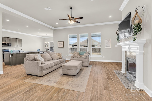 living room featuring ceiling fan with notable chandelier, a premium fireplace, baseboards, ornamental molding, and light wood finished floors