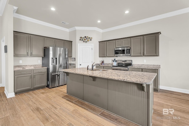 kitchen with light stone counters, stainless steel appliances, ornamental molding, light wood-type flooring, and a center island with sink