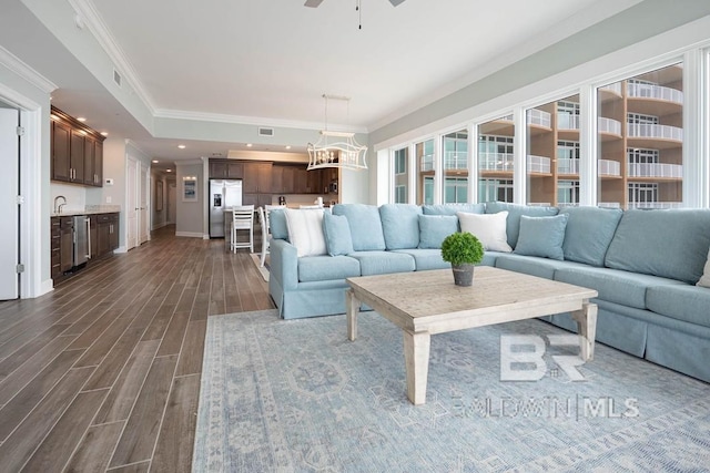 living room featuring dark wood-style flooring, visible vents, crown molding, baseboards, and ceiling fan with notable chandelier