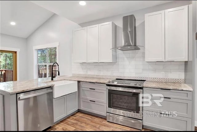 kitchen with wall chimney range hood, sink, stainless steel appliances, light stone counters, and kitchen peninsula