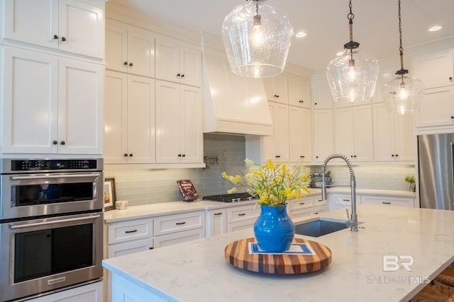 kitchen featuring custom range hood, appliances with stainless steel finishes, a kitchen island with sink, a sink, and white cabinetry