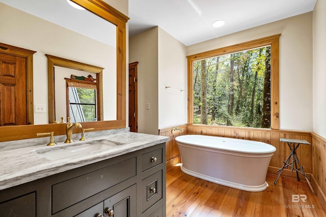 full bathroom featuring vanity, wood walls, wood-type flooring, a soaking tub, and a wainscoted wall