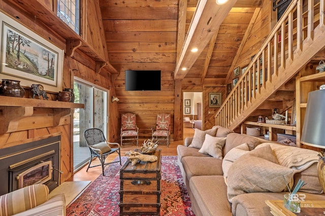 living room featuring beamed ceiling, wooden walls, wood finished floors, stairway, and a glass covered fireplace