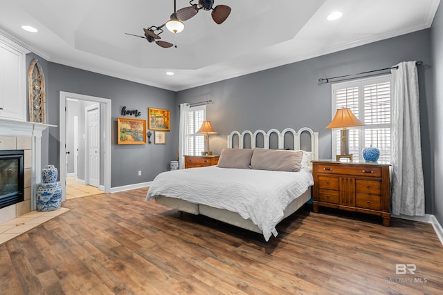 bedroom featuring a tiled fireplace, a raised ceiling, ceiling fan, hardwood / wood-style flooring, and ornamental molding