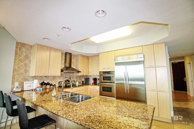 kitchen with wall chimney range hood, sink, light brown cabinetry, appliances with stainless steel finishes, and kitchen peninsula