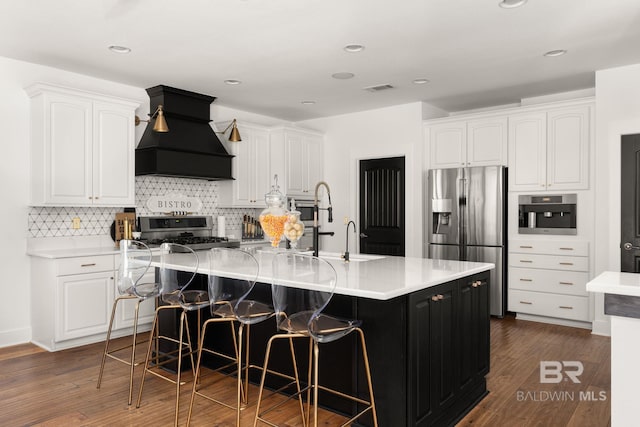 kitchen featuring premium range hood, a kitchen island with sink, appliances with stainless steel finishes, white cabinets, and dark hardwood / wood-style flooring