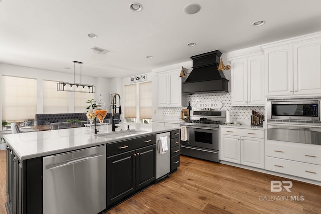 kitchen featuring white cabinetry, hanging light fixtures, stainless steel appliances, custom range hood, and an island with sink