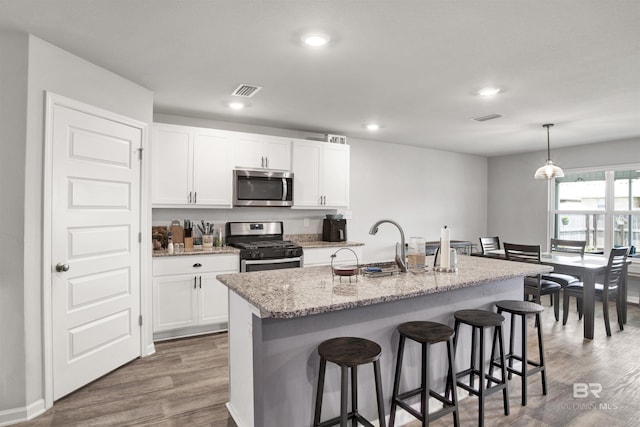 kitchen featuring appliances with stainless steel finishes, a center island with sink, decorative light fixtures, hardwood / wood-style floors, and white cabinetry