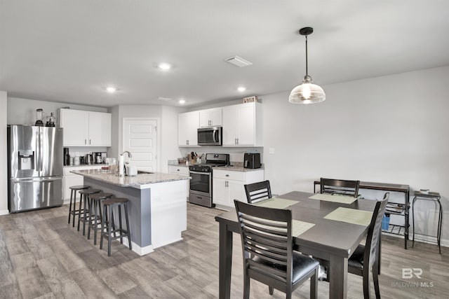 kitchen with light stone counters, stainless steel appliances, light hardwood / wood-style floors, white cabinetry, and an island with sink