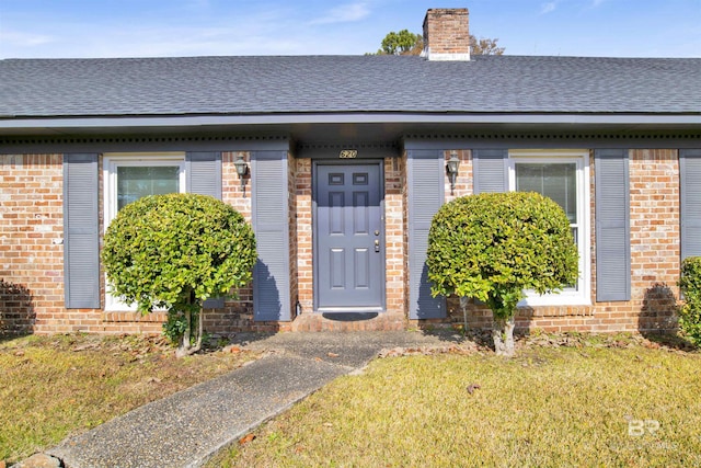 entrance to property featuring brick siding, a shingled roof, a chimney, and a lawn