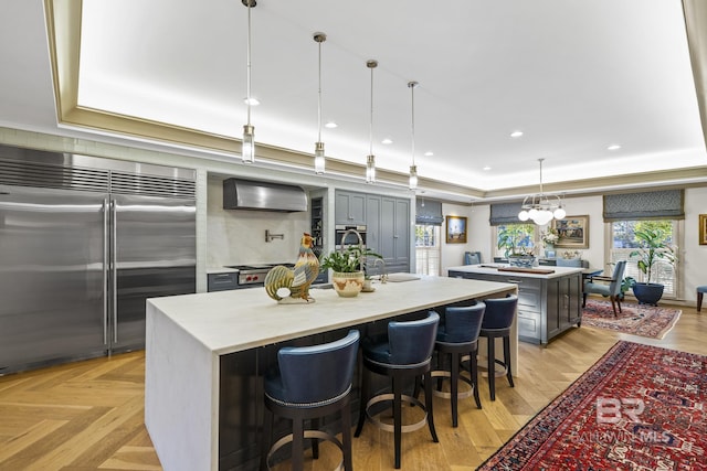 kitchen with stainless steel appliances, gray cabinets, wall chimney exhaust hood, a raised ceiling, and a center island with sink