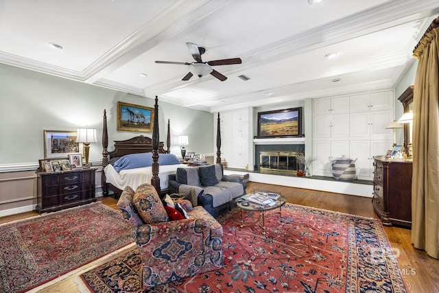 bedroom featuring a wainscoted wall, wood finished floors, ornamental molding, beam ceiling, and a glass covered fireplace