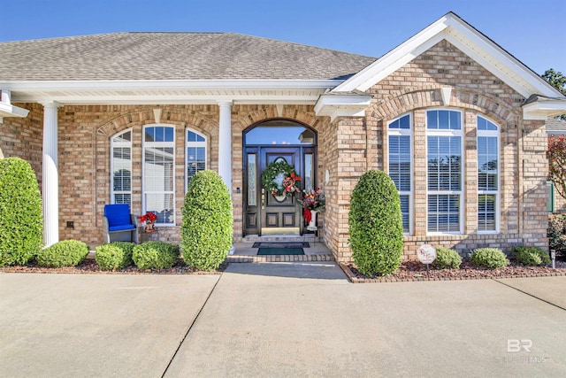 view of exterior entry with brick siding, roof with shingles, and a porch