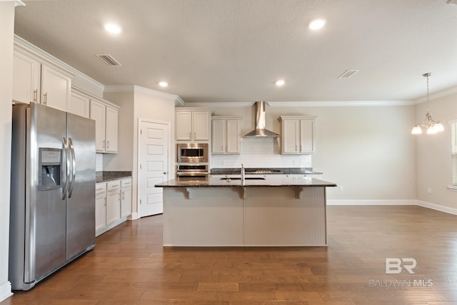 kitchen with wall chimney exhaust hood, appliances with stainless steel finishes, dark stone countertops, and a kitchen island with sink