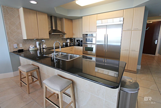 kitchen with light brown cabinetry, appliances with stainless steel finishes, wall chimney range hood, and backsplash