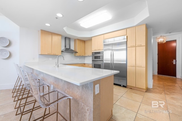 kitchen with a peninsula, wall chimney exhaust hood, light brown cabinets, and appliances with stainless steel finishes