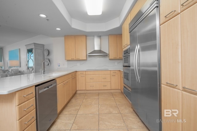 kitchen featuring light brown cabinets, a peninsula, a sink, stainless steel appliances, and wall chimney exhaust hood