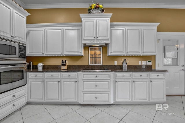 kitchen featuring appliances with stainless steel finishes, light tile flooring, dark stone countertops, white cabinetry, and crown molding