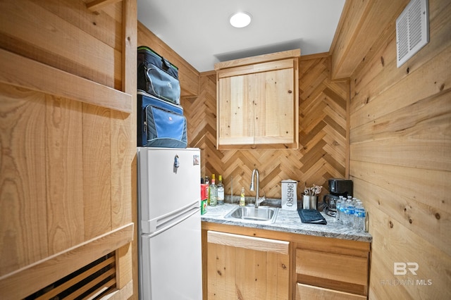 kitchen with a sink, freestanding refrigerator, light stone countertops, wooden walls, and light brown cabinets