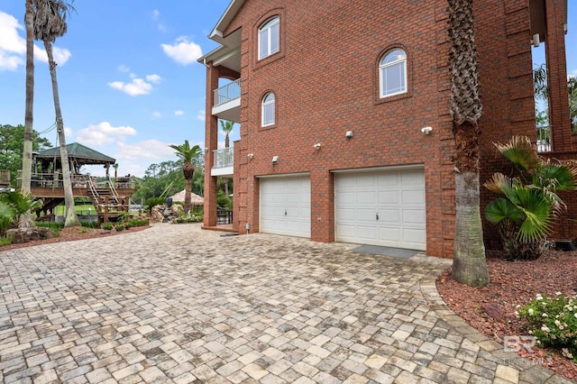 view of side of property with an attached garage, a balcony, decorative driveway, a gazebo, and brick siding