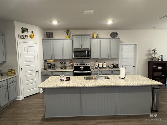 kitchen with gray cabinets, light stone counters, a kitchen island with sink, and appliances with stainless steel finishes