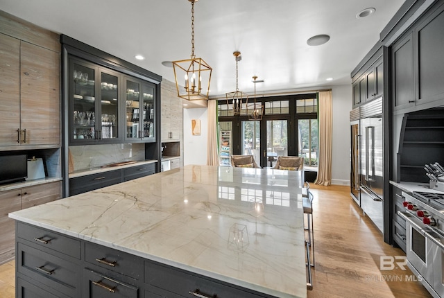 kitchen featuring hanging light fixtures, high end stainless steel range, light stone countertops, light wood finished floors, and glass insert cabinets