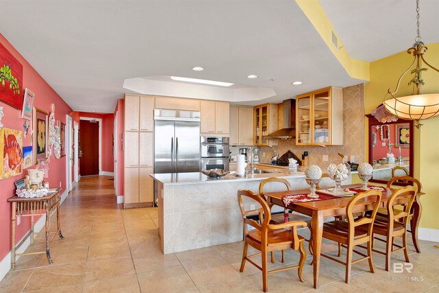 kitchen with wall chimney range hood, kitchen peninsula, backsplash, appliances with stainless steel finishes, and light brown cabinetry