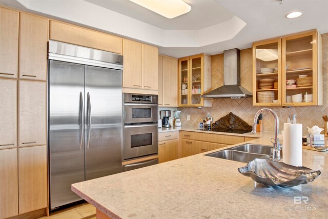 kitchen with wall chimney range hood, decorative backsplash, a raised ceiling, light brown cabinetry, and stainless steel appliances