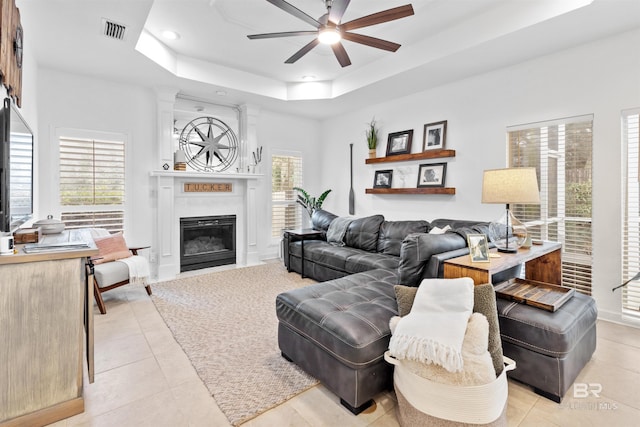 living room with a tray ceiling, light tile patterned floors, visible vents, a ceiling fan, and a glass covered fireplace