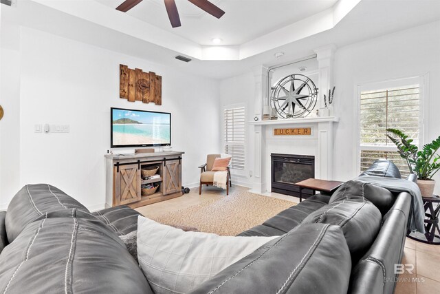 living area with a tray ceiling, visible vents, a fireplace with flush hearth, ceiling fan, and baseboards