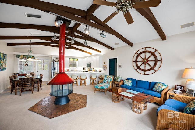 living area with a ceiling fan, a wood stove, visible vents, and vaulted ceiling with beams