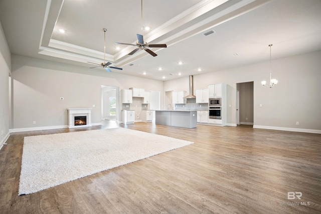 unfurnished living room featuring a raised ceiling, ceiling fan with notable chandelier, ornamental molding, and hardwood / wood-style flooring