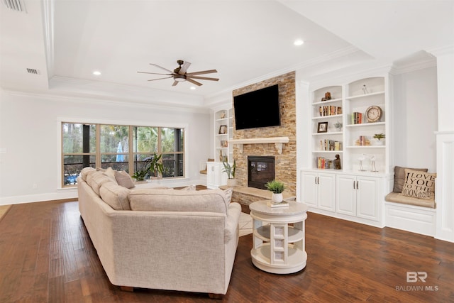 living room featuring a stone fireplace, ceiling fan, crown molding, dark wood-type flooring, and built in shelves