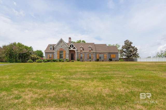 tudor-style house featuring a chimney, fence, a front yard, and stone siding