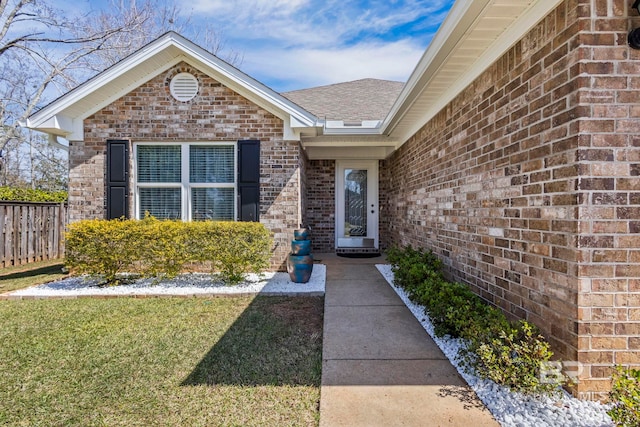 doorway to property featuring a yard, brick siding, roof with shingles, and fence