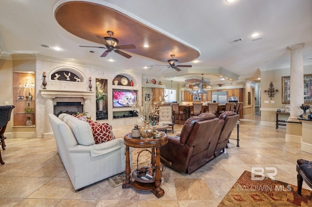 living room featuring decorative columns, crown molding, a large fireplace, and ceiling fan