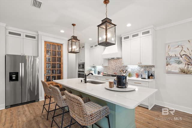 kitchen with stainless steel appliances, an island with sink, premium range hood, pendant lighting, and sink