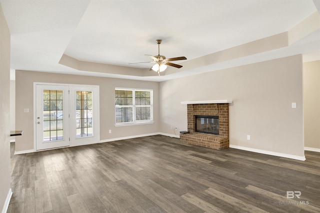 unfurnished living room with a brick fireplace, ceiling fan, baseboards, a raised ceiling, and dark wood-style flooring