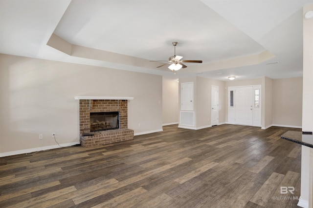 unfurnished living room with a fireplace, a tray ceiling, dark wood-style floors, and baseboards