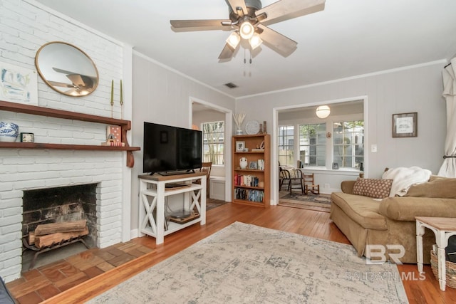 living area with wood finished floors, visible vents, ceiling fan, crown molding, and a brick fireplace