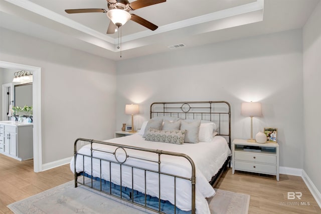 bedroom with ceiling fan, ensuite bath, light wood-type flooring, and a tray ceiling