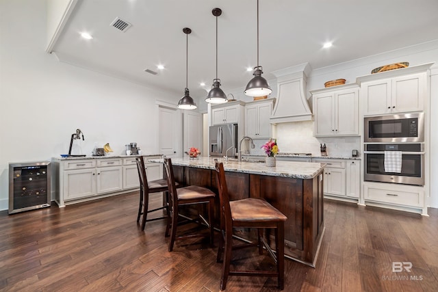 kitchen featuring appliances with stainless steel finishes, decorative light fixtures, white cabinetry, an island with sink, and beverage cooler