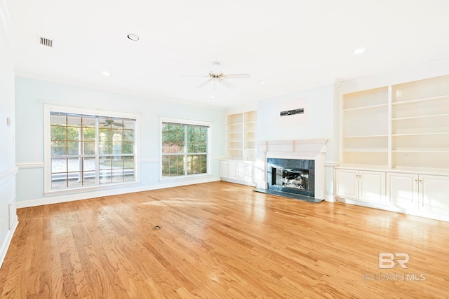 unfurnished living room featuring crown molding, built in features, a premium fireplace, ceiling fan, and light wood-type flooring